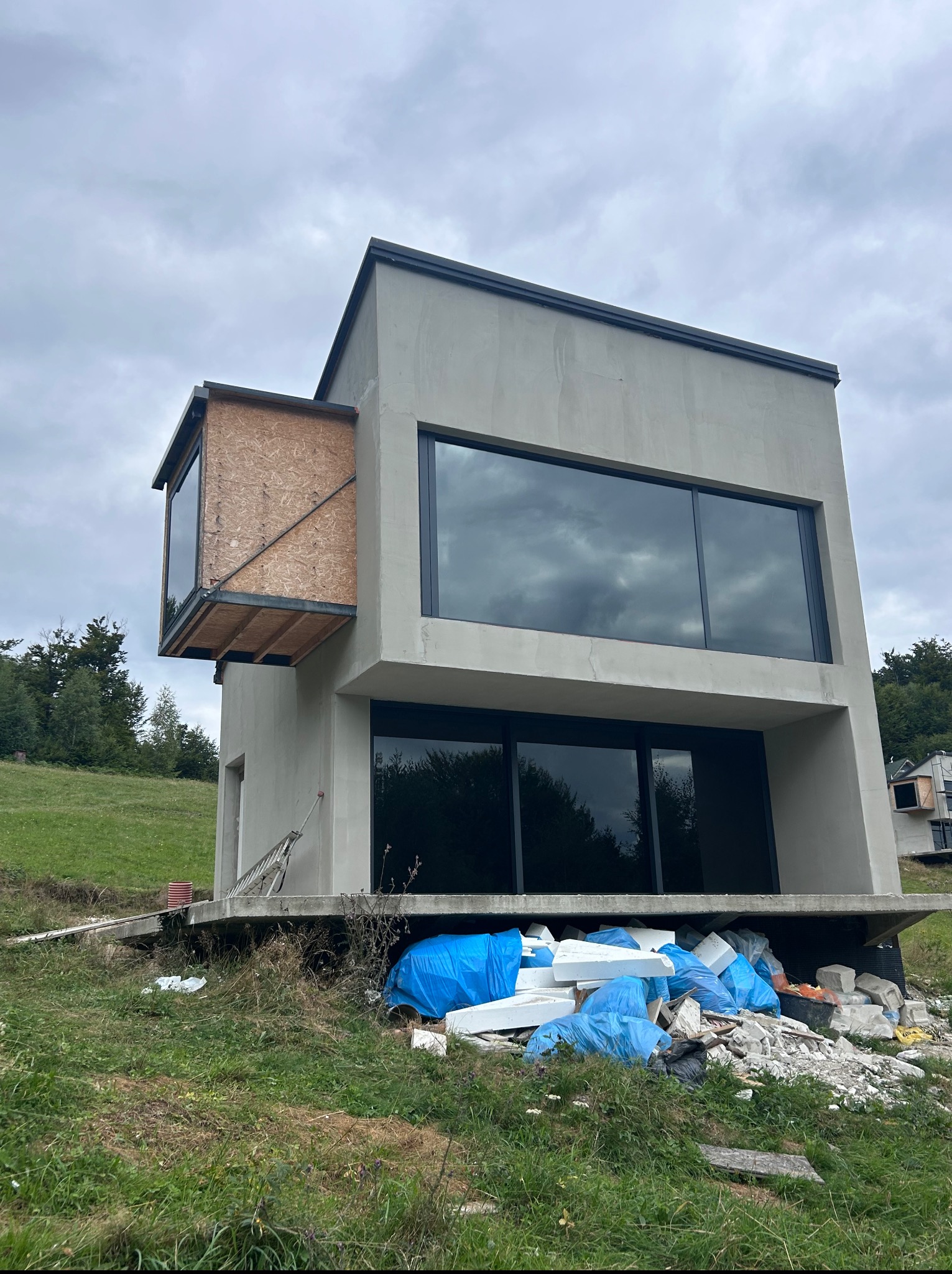 Surowy, dwukondygnacyjny dom z dużymi oknami i dobudówką z OSB, w trakcie budowy, z materiałami budowlanymi na zewnątrz. Widok na zielone wzgórze i pochmurne niebo.