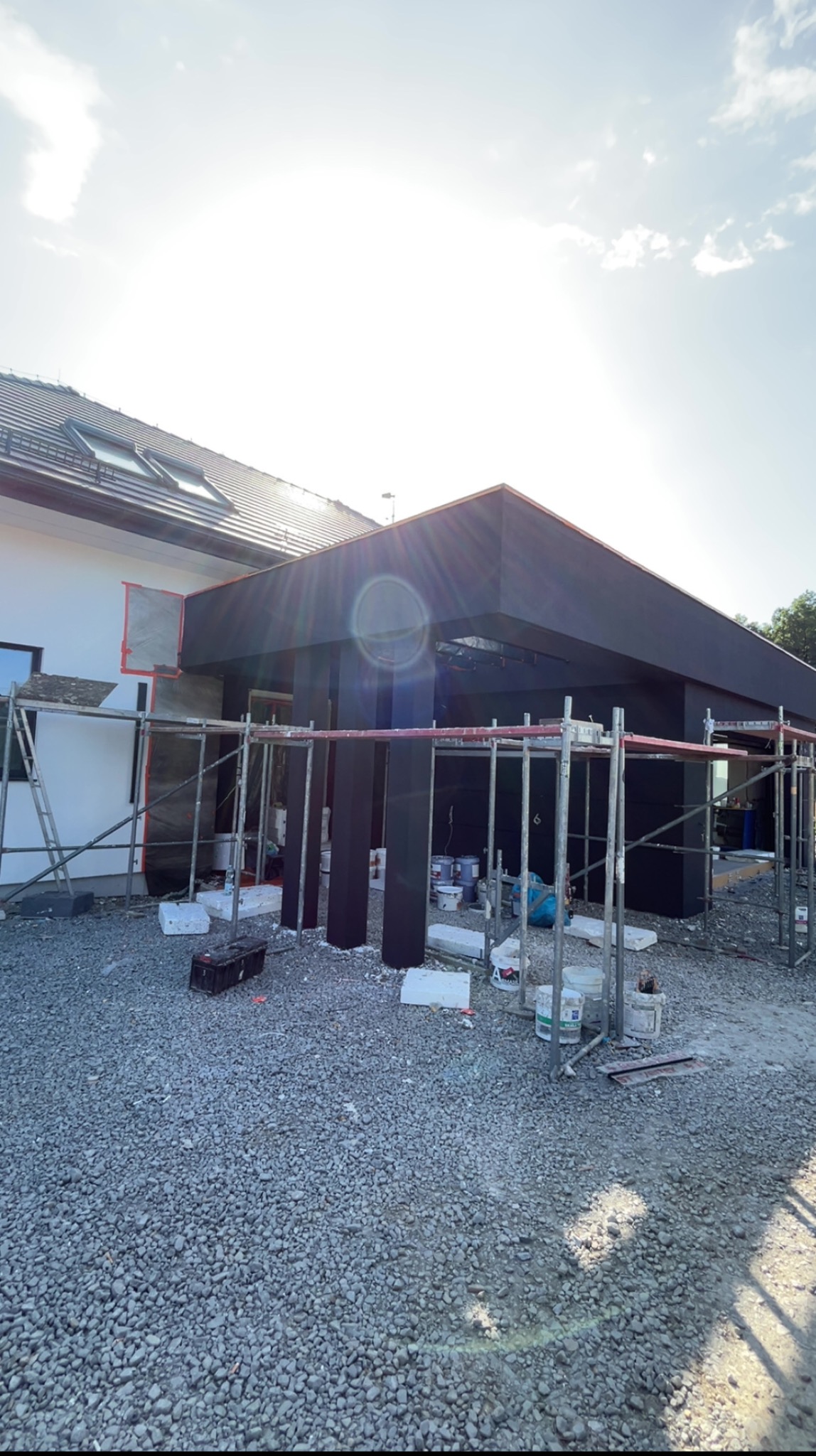 Dom w trakcie wykończenia elewacji: czarne elementy konstrukcyjne w rusztowaniach, przygotowania do prac na ścianie, gruz na podłożu. Słoneczny dzień.