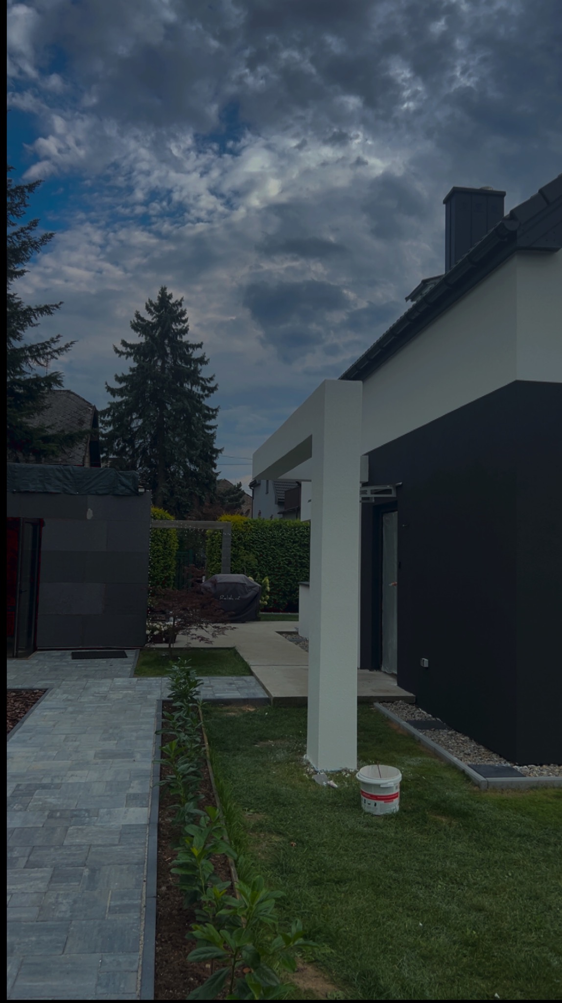 Nowoczesny dom z antracytową elewacją i jasnymi elementami architektonicznymi. Widoczny fragment ogrodu z kostką brukową i świeżo posadzoną zielenią.