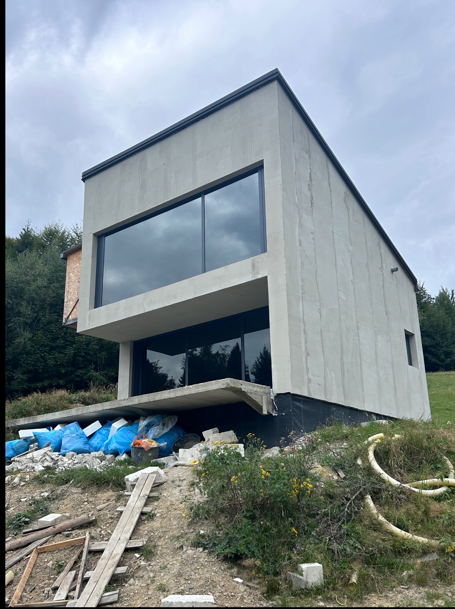 Surowy, minimalistyczny budynek z dużymi oknami w trakcie prac wykończeniowych, z widocznym ociepleniem i elementami konstrukcyjnymi w otoczeniu zieleni.