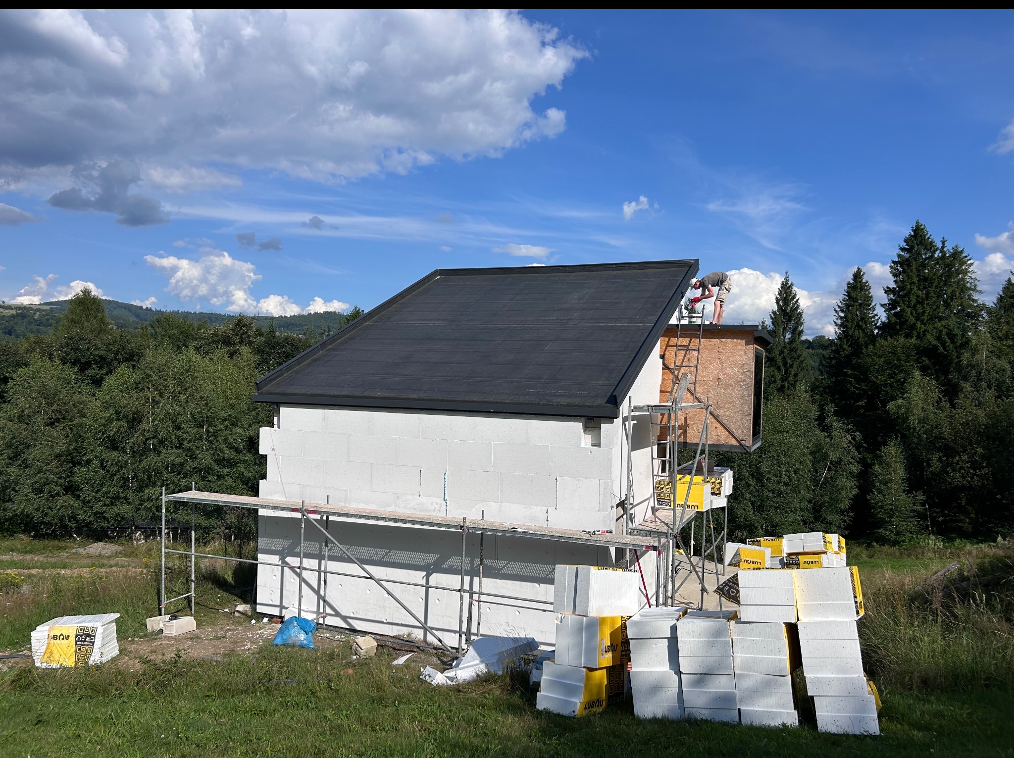 Budowa domu jednorodzinnego w trakcie ocieplania, widoczne rusztowania, białe bloczki, ocieplenie i pracownik na dachu na tle zieleni i błękitnego nieba.