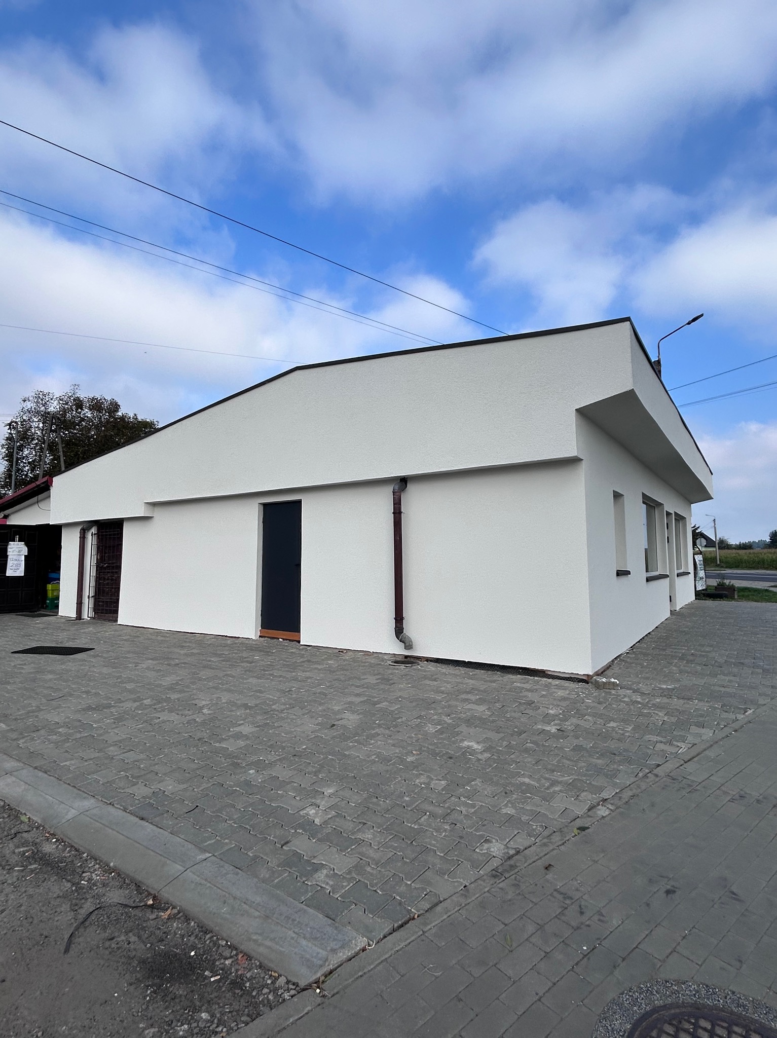 Biały budynek po remoncie z ciemnymi drzwiami i rynną na tle błękitnego nieba z chmurami i szarej kostki brukowej. Widoczny fragment ulicy i lampa.