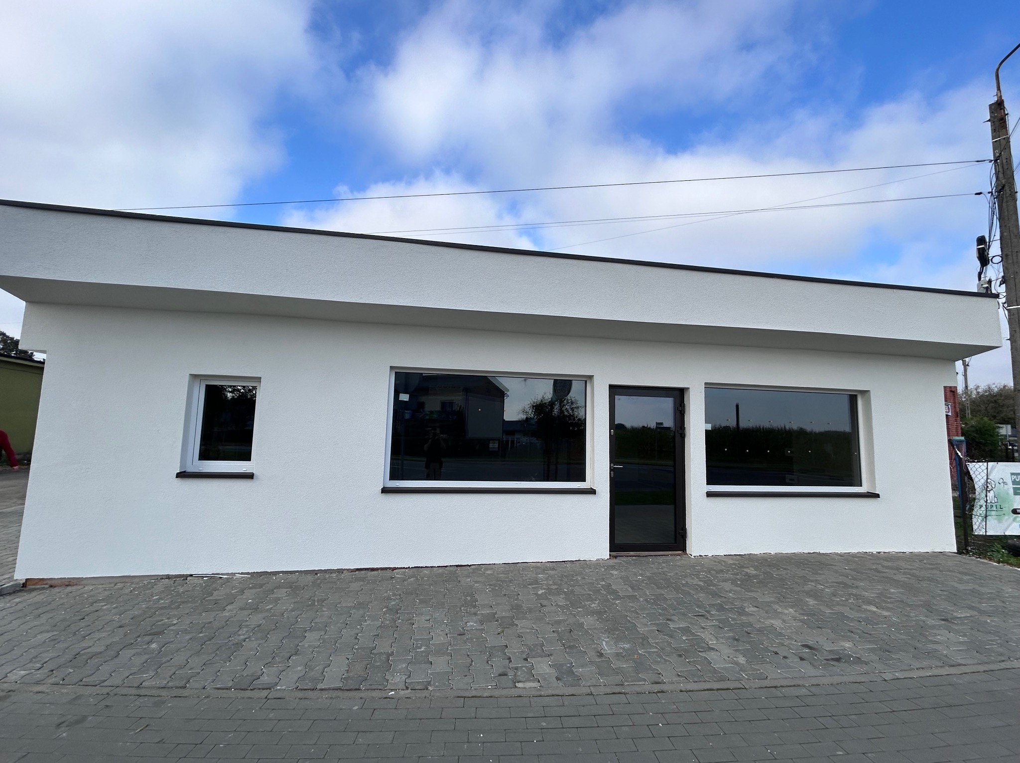 Biały, jednopiętrowy budynek z ciemnymi oknami i drzwiami, wykończony z zewnątrz. Ułożona kostka brukowa przed budynkiem. Błękitne niebo z chmurami w tle.