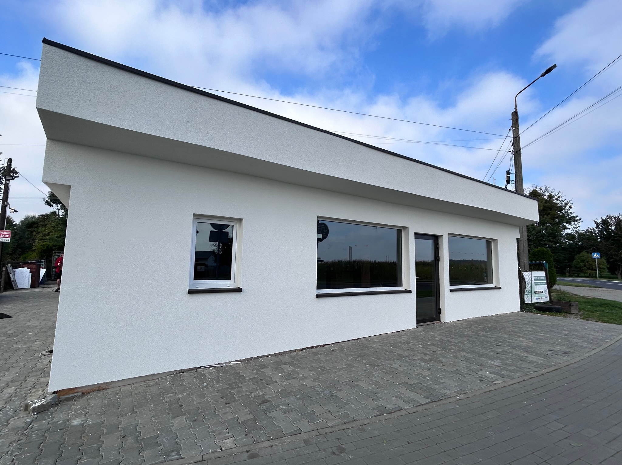 Biały budynek z oknami i czarnym dachem na tle błękitnego nieba. Widoczny chodnik z kostki brukowej i fragment ulicy. Minimalistyczna architektura budynku.