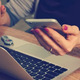 Kobieta używa smartfona przy laptopie, na biurku leży token USB. Dłonie z fuksjowym manicure i złotym pierścionkiem. Praca zdalna, e-administracja, bezpieczeństwo danych.