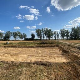 Uslugi koparko-ładowarką - Przygotowany teren pod budowę, widoczne ślady po maszynach, wyznaczony obszar, w tle drzewa i błękitne niebo z chmurami.