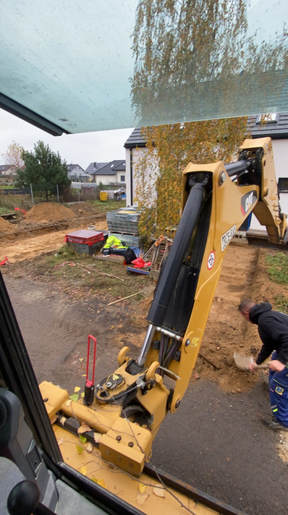 Żółta koparka CAT w trakcie prac ziemnych przy budowie domu jednorodzinnego. Widok z kabiny operatora, mężczyzna z łopatą wyrównuje wykop.