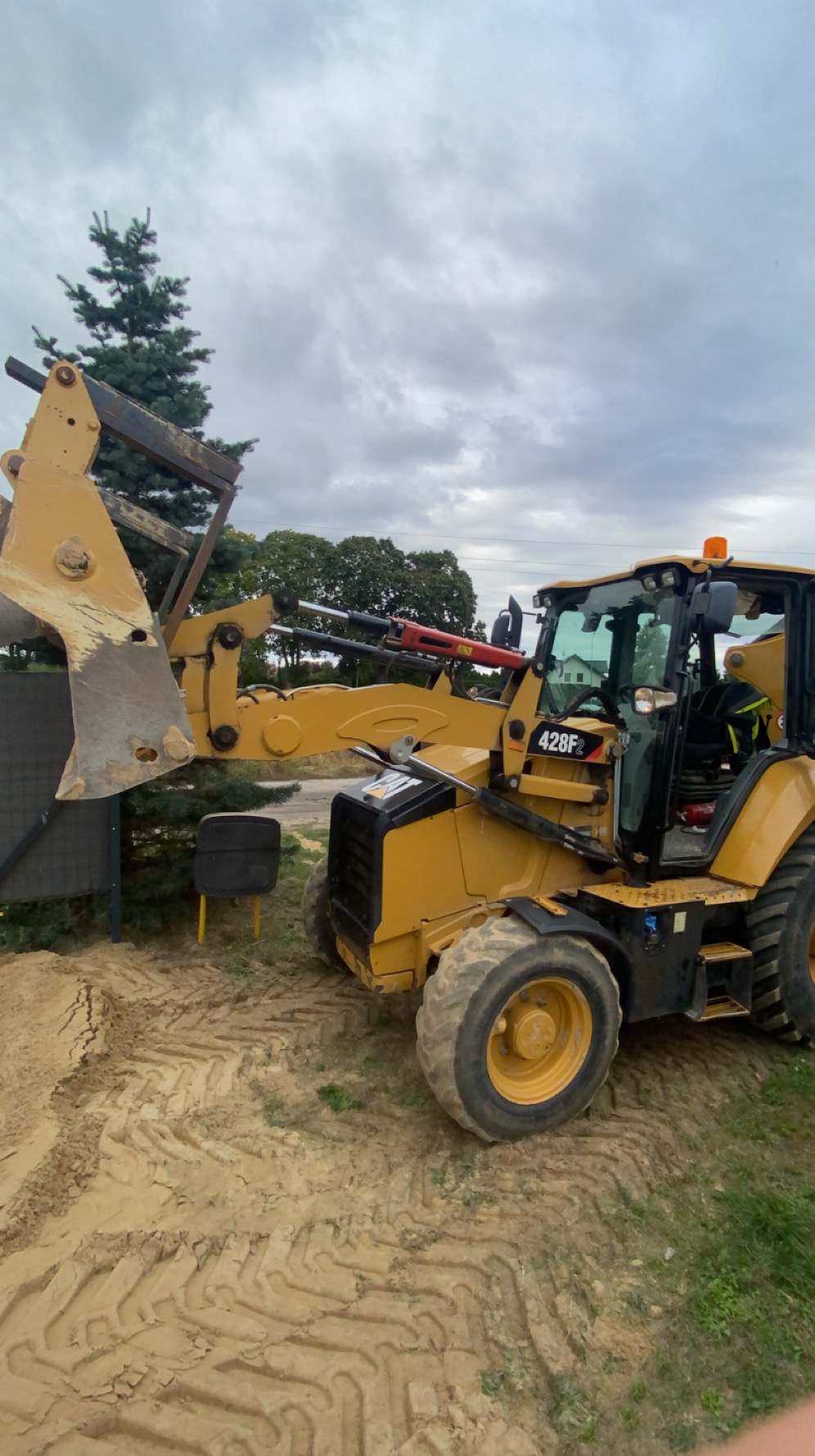 Żółta koparko-ładowarka CAT 428F2 na placu budowy, widoczna z boku, z łyżką skierowaną w lewo, na tle zieleni i pochmurnego nieba.