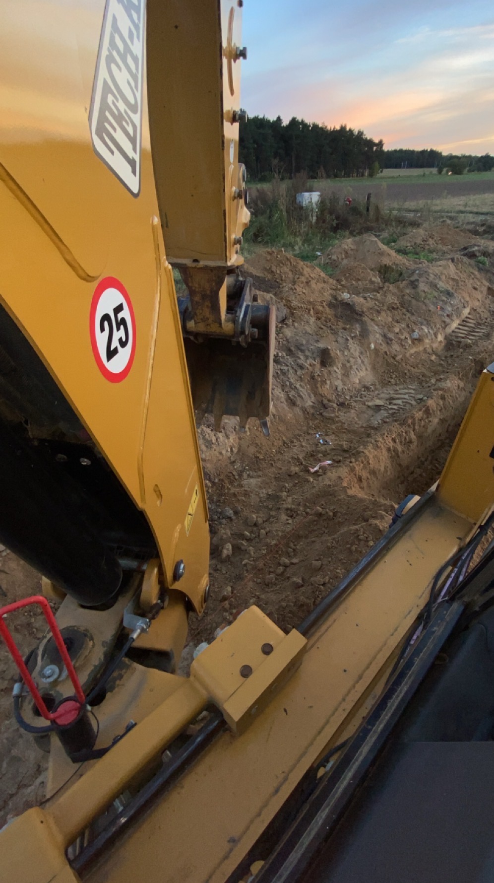 Żółta koparka w trakcie pracy przy wykopie, w tle widoczne pole i las, oświetlenie wieczorne. Na ramieniu koparki naklejka z ograniczeniem prędkości do 25 km/h.