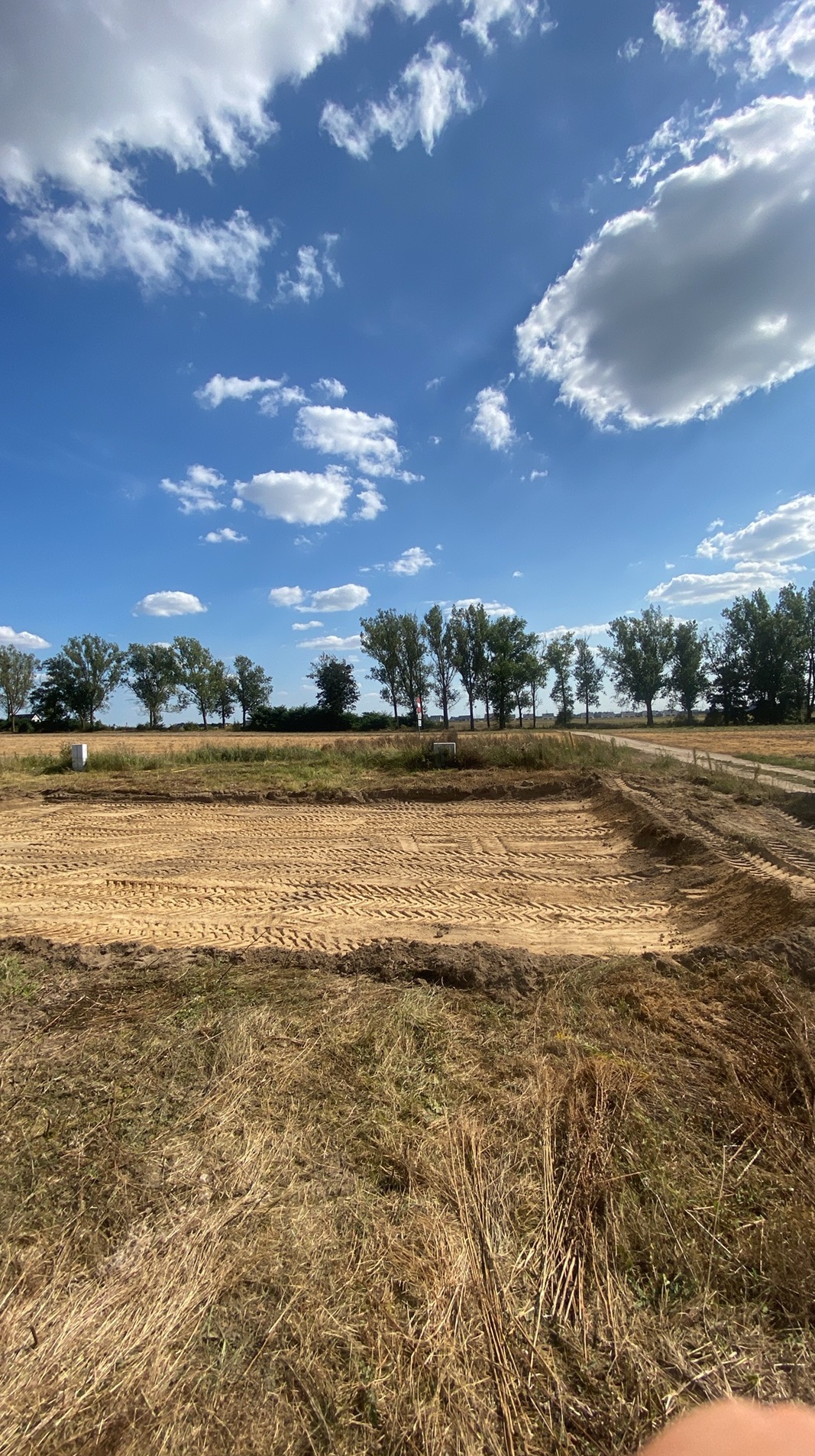 Przygotowany teren pod budowę, widoczne ślady po maszynach, wyznaczony obszar, w tle drzewa i błękitne niebo z chmurami.