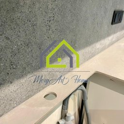 MeryArt Home - Szara ściana z gniazdkiem nad beżowym blatem z otworem i szarą instalacją hydrauliczną w wykończonej łazience.