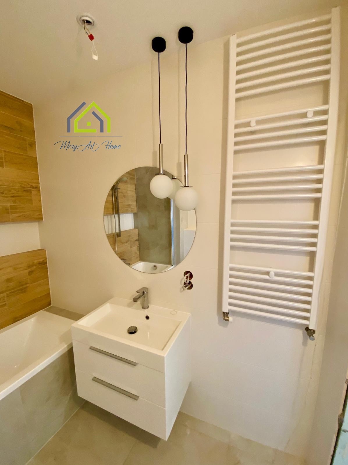 Jasna łazienka po remoncie: biała umywalka z szafką, okrągłe lustro, grzejnik drabinkowy i wiszące lampy. Fragment wanny z drewnopodobnymi płytkami.