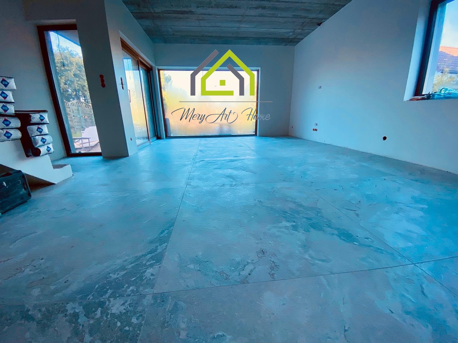 Surowe wnętrze w trakcie wykończenia: wylewka podłogowa, ściany przygotowane do malowania, duże okna. Widoczne puszki elektryczne. W tle materiały budowlane.