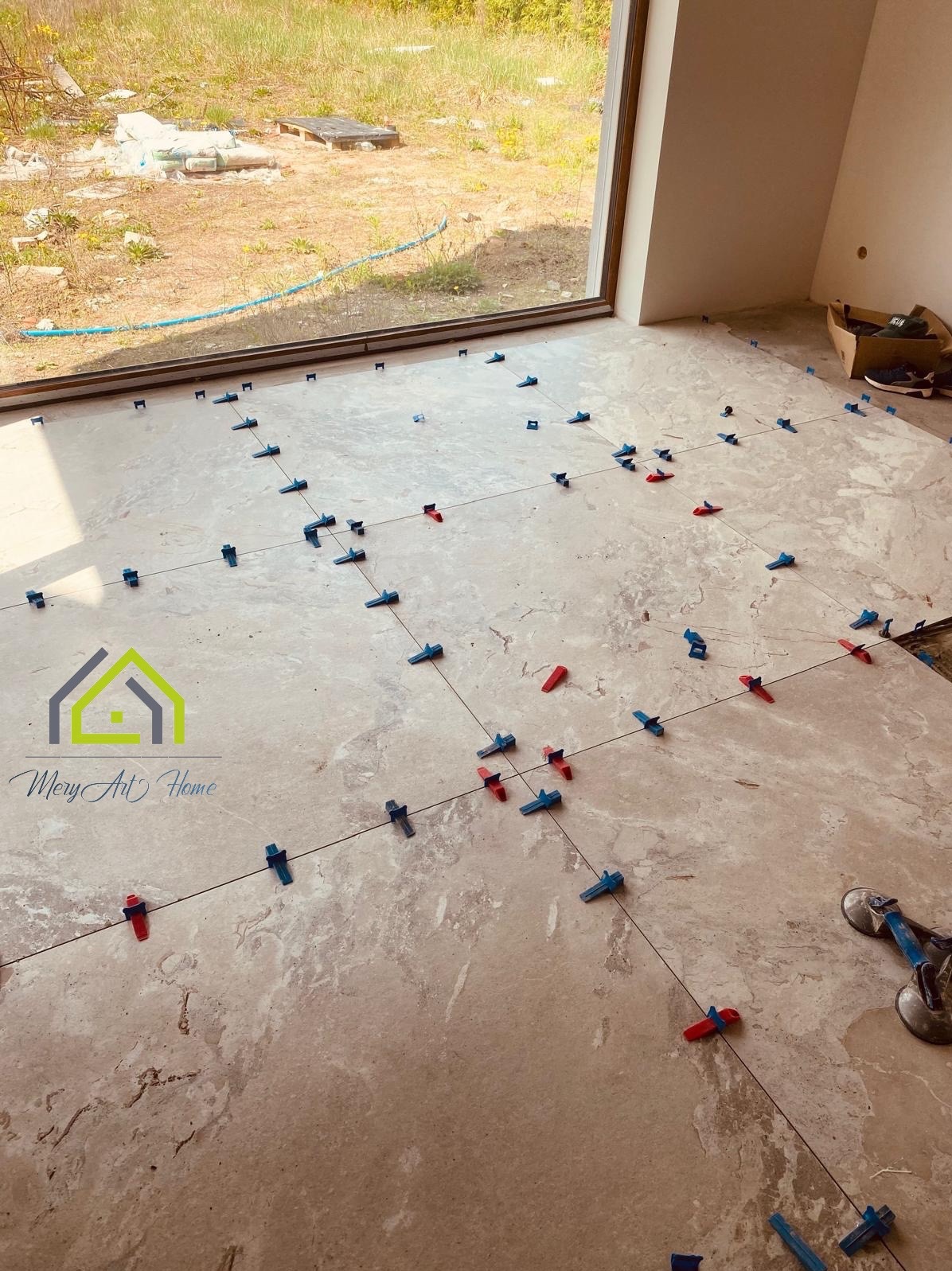 Jasne płytki podłogowe w trakcie układania, z użyciem poziomych i pionowych linek oraz klipsów. Widoczne przyssawki do przenoszenia płytek i logo firmy.