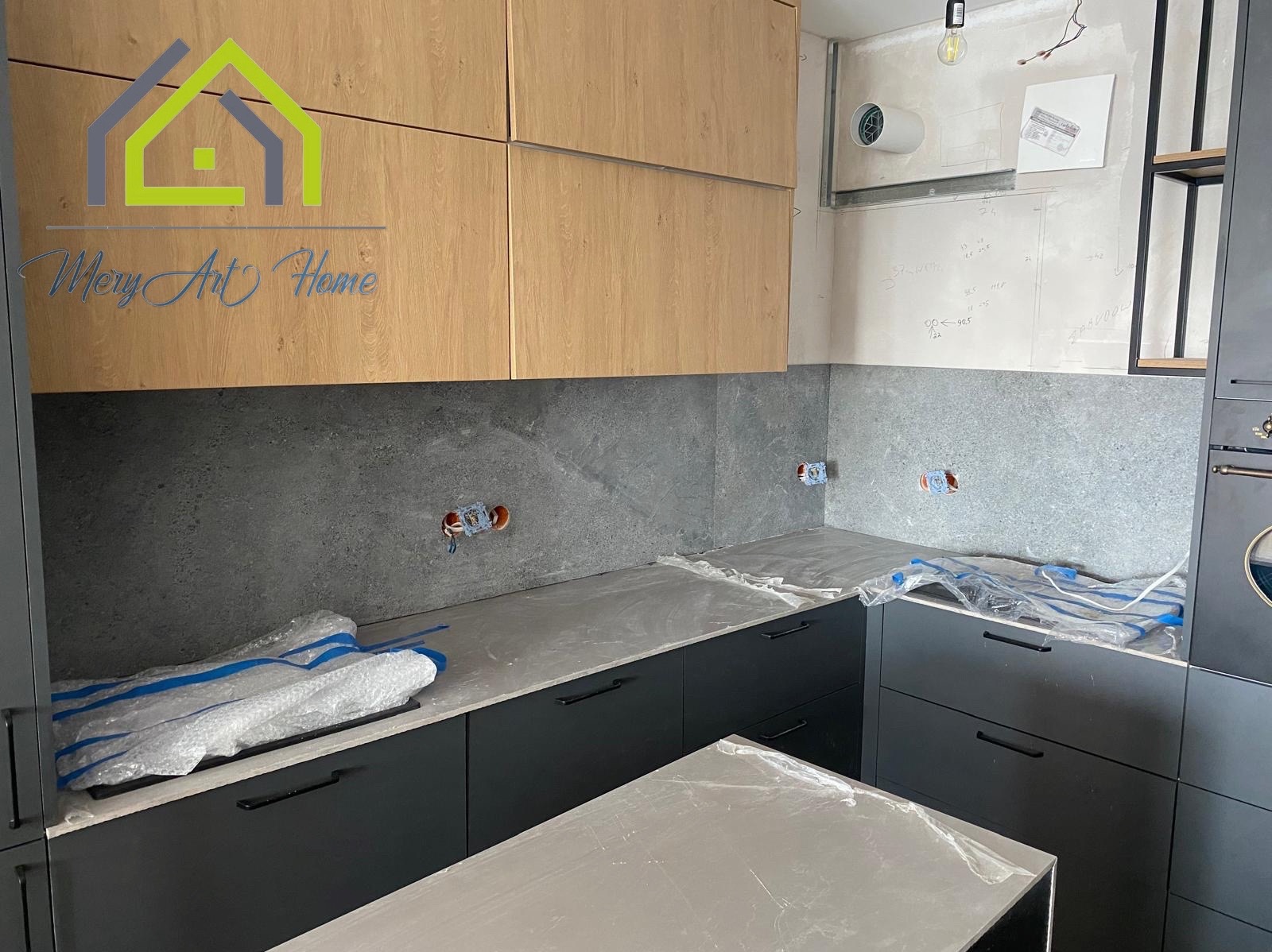 Częściowo wykończona kuchnia z drewnianymi szafkami, szarym blatem i płytkami. Widoczne gniazdka elektryczne i zabezpieczone folią blaty. Ściana z notatkami.