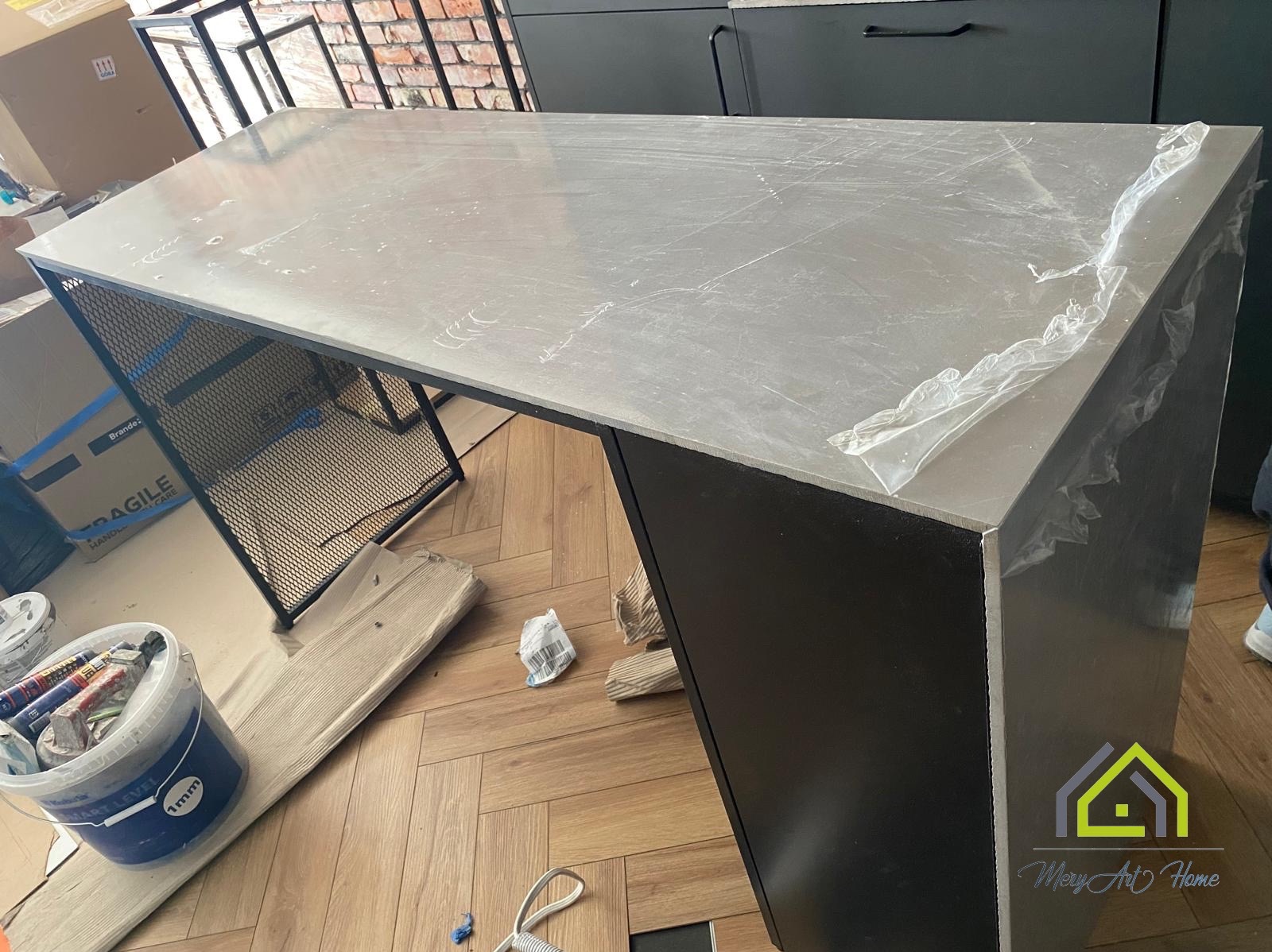 Nowoczesna wyspa kuchenna z szarą, zabezpieczoną folią płytą i czarną podstawą na parkiecie, z widocznymi akcesoriami malarskimi w tle.
