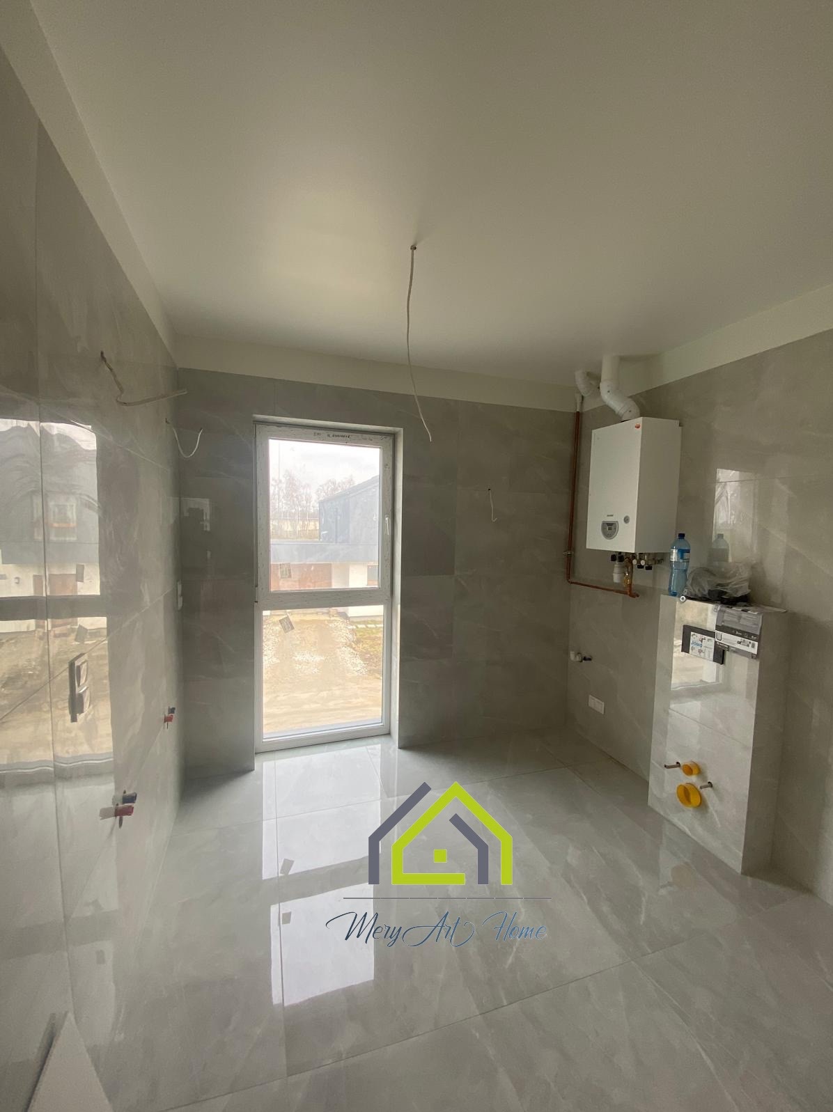 Jasna łazienka po remoncie: szare płytki na ścianach i podłodze, biały bojler i zabudowa WC. Widoczne wystające kable i rury. Okno z widokiem na zewnątrz.