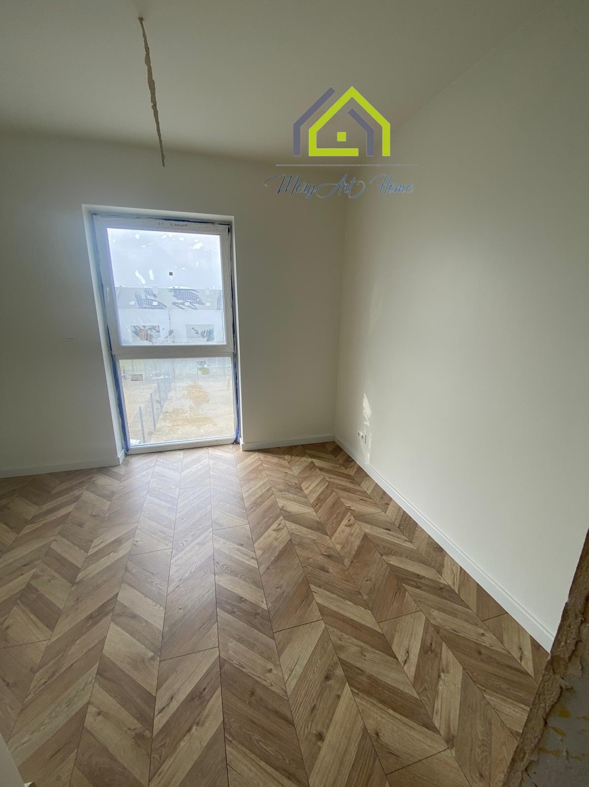 Jasne panele ułożone w jodełkę w pustym pokoju z oknem i białymi ścianami. Widoczny fragment surowego wykończenia ściany.