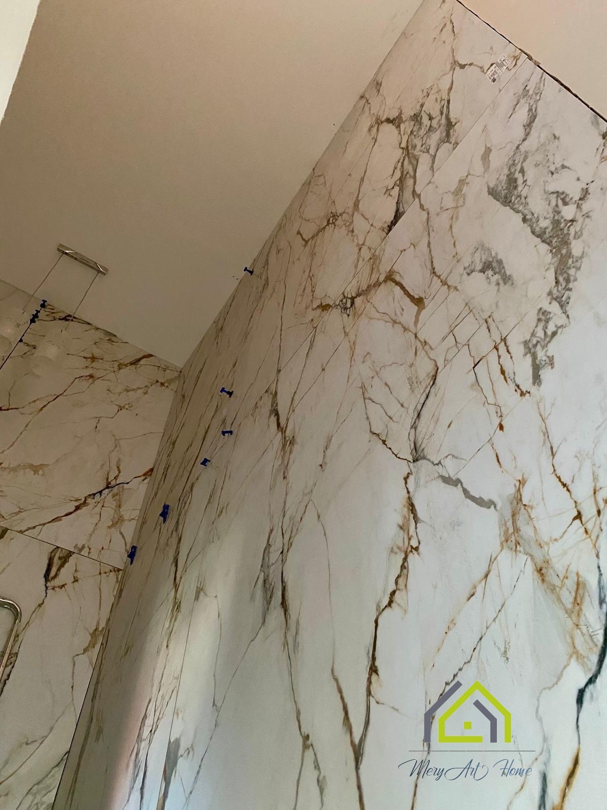 Białe płytki imitujące marmur z brązowymi żyłami na ścianie łazienki, z niebieskimi krzyżykami dystansowymi, widok z dołu.