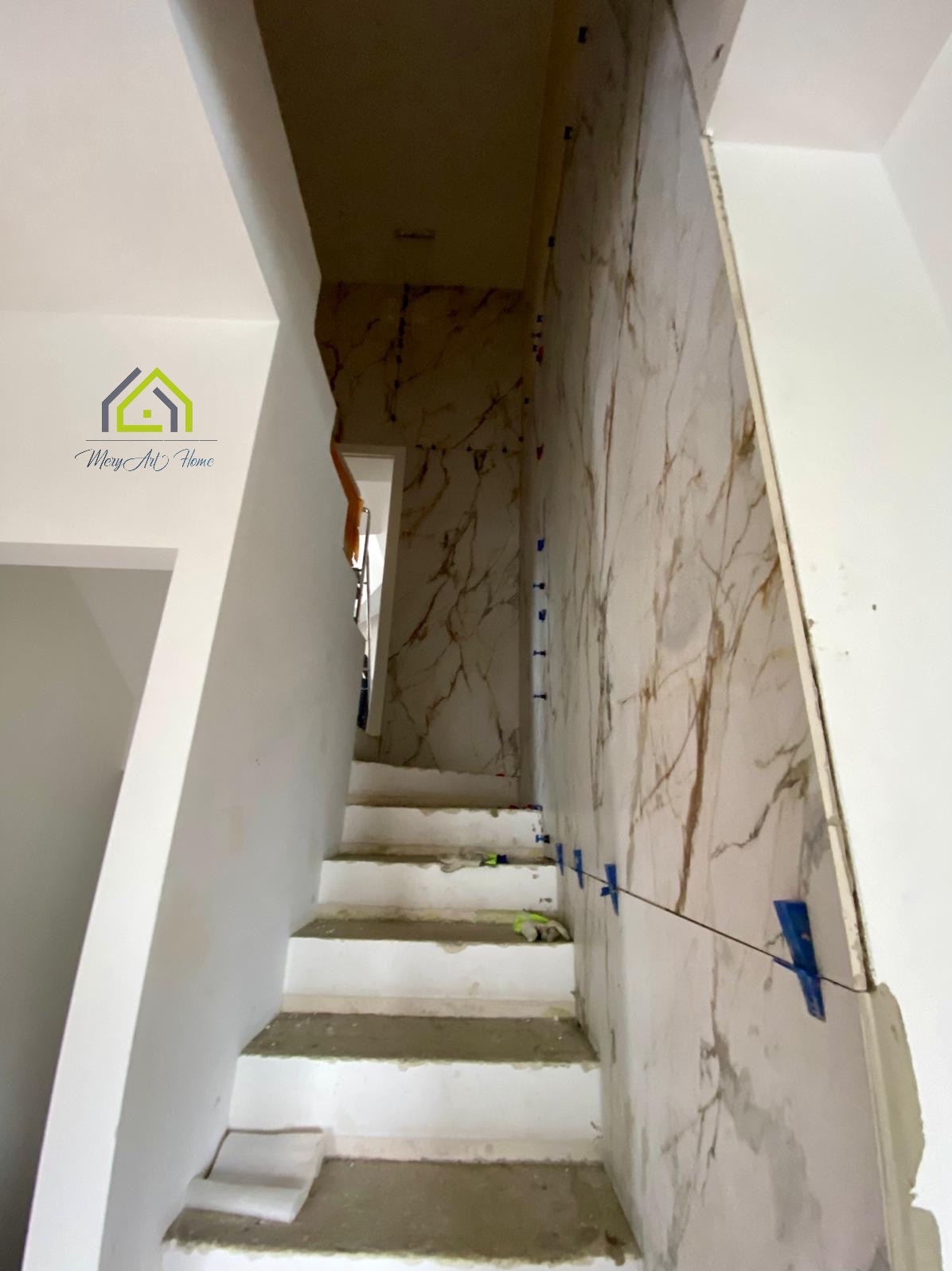 Schody w trakcie wykańczania z marmurowymi płytkami na ścianie, widoczne niebieskie krzyżyki dystansowe, perspektywa od dołu.