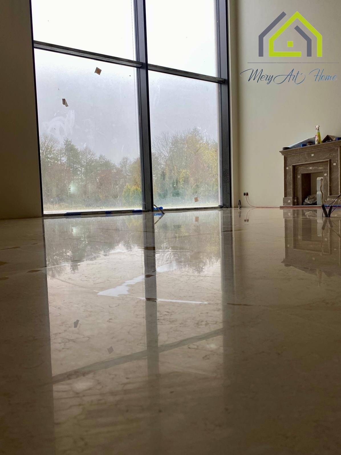 Błyszcząca, jasna marmurowa podłoga w przestronnym wnętrzu z dużym oknem i dekoracyjnym kominkiem. Widoczne ślady po pracach wykończeniowych.