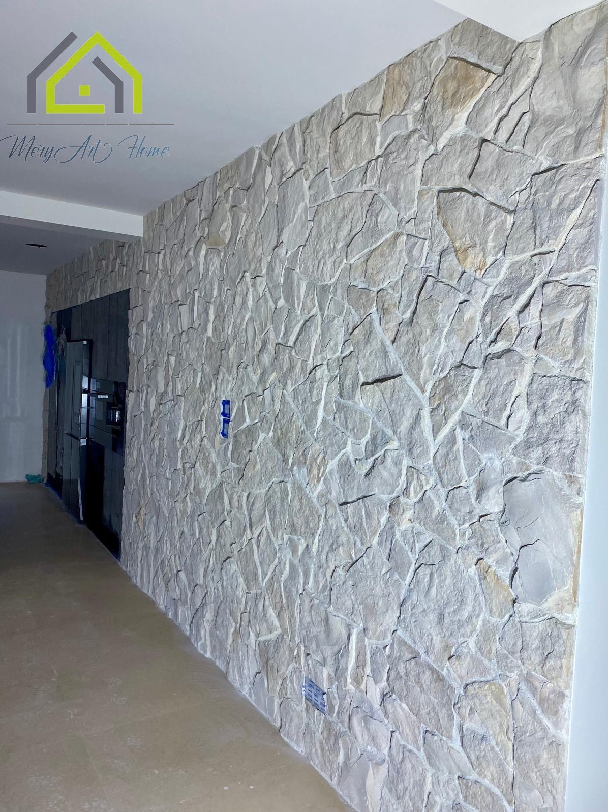 Ściana wykończona dekoracyjnym kamieniem w odcieniach beżu i szarości. Widoczne gniazdka elektryczne. W tle fragment kuchni z czarną zabudową.