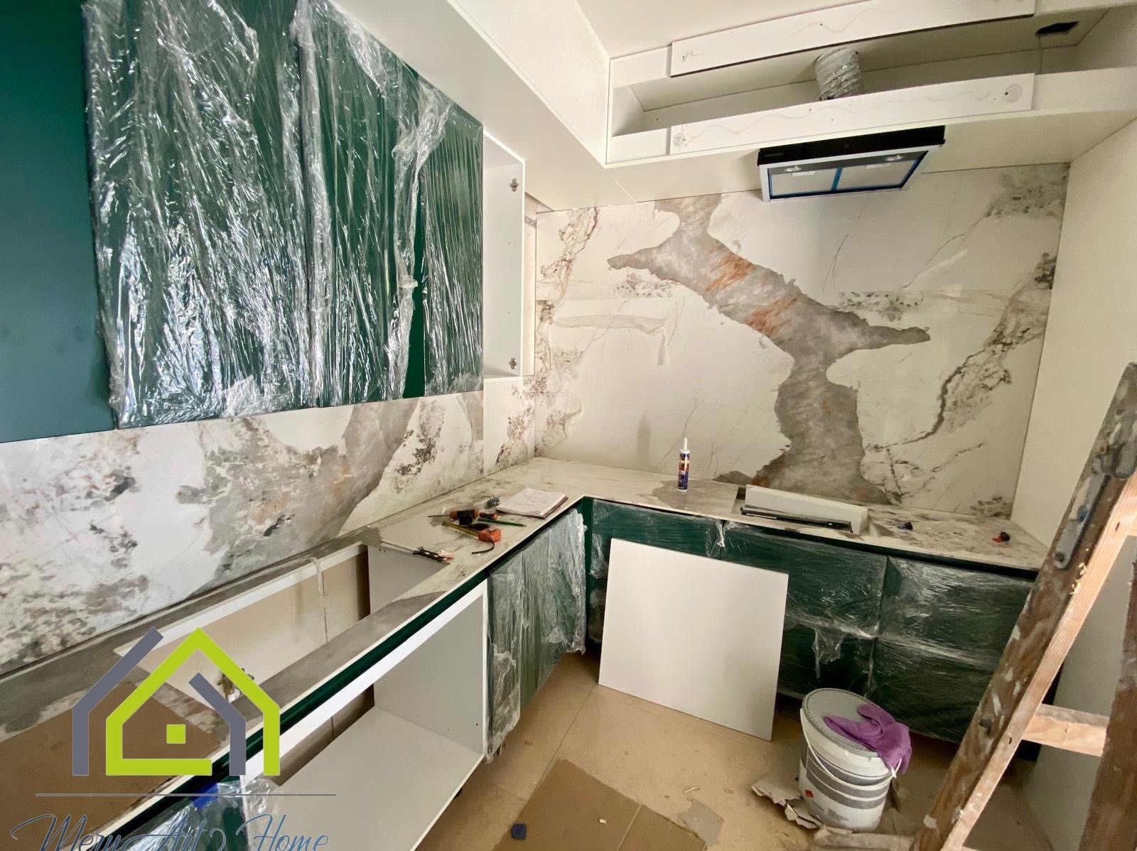 Montaż kuchni: zielone szafki częściowo owinięte folią ochronną, marmurowy backsplash, drabina i narzędzia sugerują trwający remont.