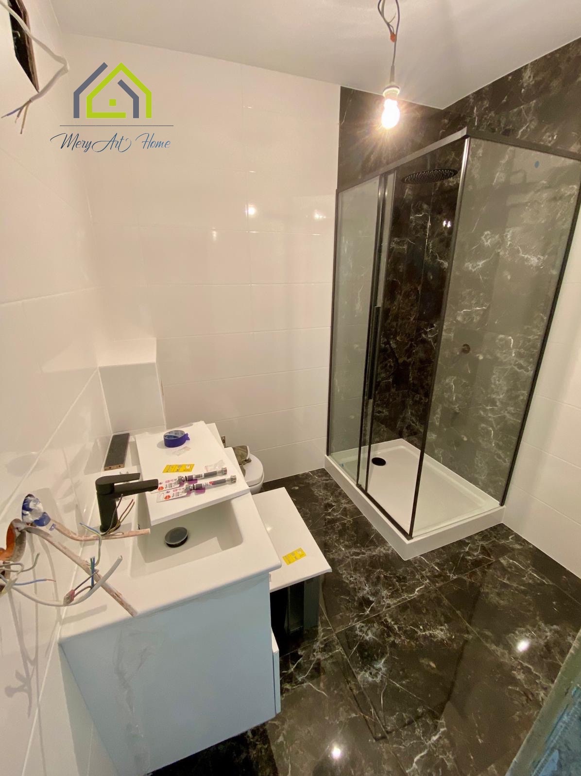 Nowoczesna łazienka z czarnymi marmurowymi płytkami podłogowymi i kabiną prysznicową. Białe płytki ścienne, umywalka w trakcie montażu. Oświetlenie sufitowe.