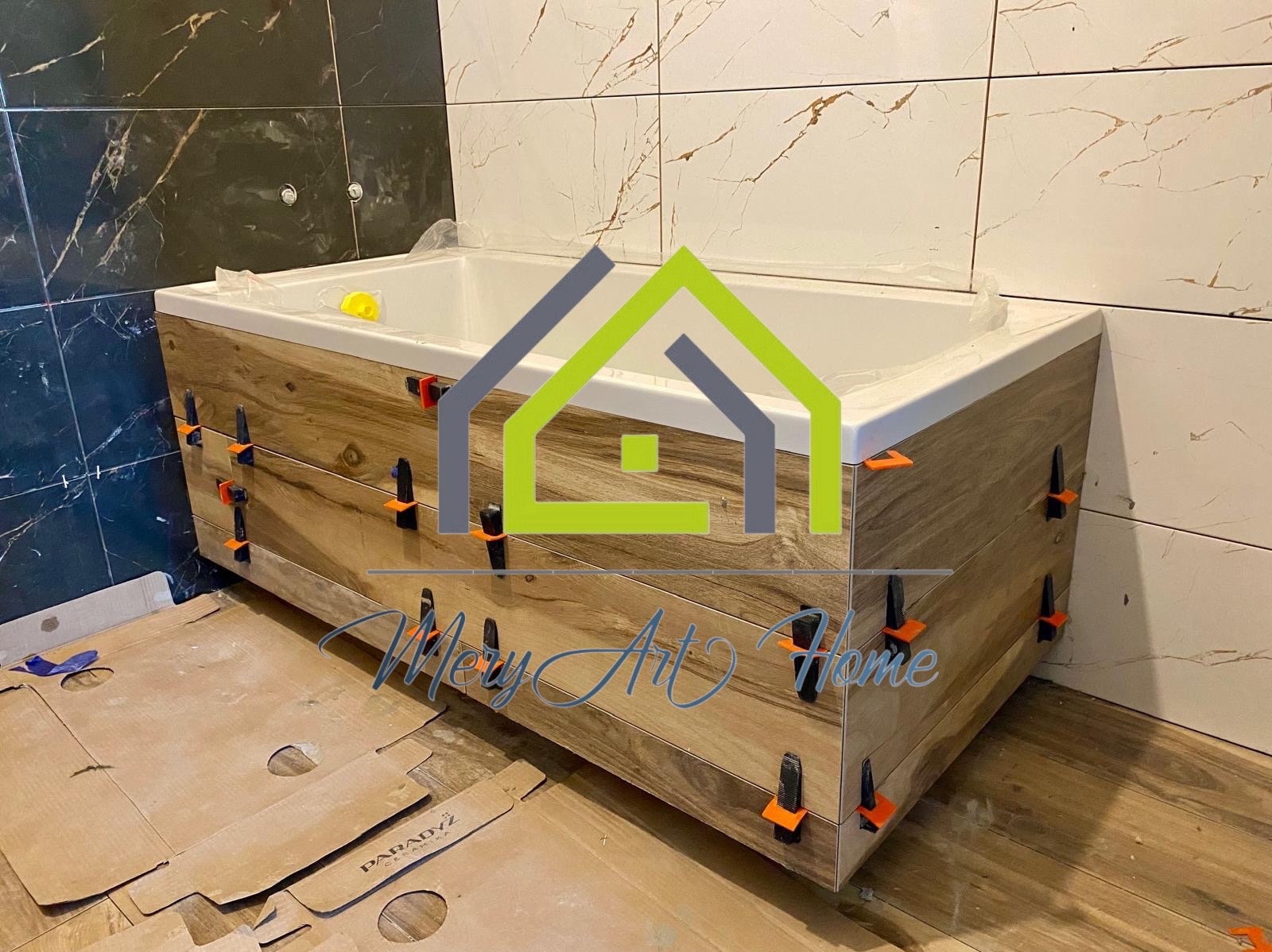 Obudowa wanny z płytek imitujących drewno, w trakcie montażu z użyciem klipsów poziomujących. W tle ściana wyłożona płytkami marmurkowymi.