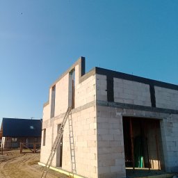 Pro-kop - Wznoszenie ścian domu z bloczków betonu komórkowego. Widoczna drabina oparta o ścianę oraz drugi budynek w tle. Stan surowy otwarty w słoneczny dzień.