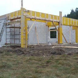 Pro-kop - Budowa domu z betonu komórkowego: ściany wzniesione, szalunki żółte, rusztowanie metalowe, drabina oparta o ścianę, widoczny samochód dostawczy na tle lasu.