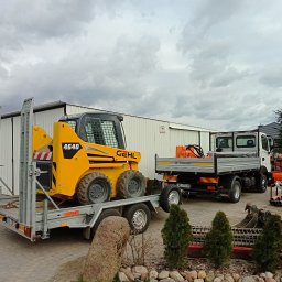 Pro-kop - Żółta ładowarka Gehl 4640 na srebrnej przyczepie, obok biała ciężarówka Pro-Kop na tle białego budynku. Elementy krajobrazu: kamienie, tuje, pochmurne niebo.