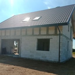 Pro-kop - Surowy dom z białych pustaków z ciemną dachówką i dwoma oknami dachowymi, w otoczeniu zieleni i błękitnego nieba. Widoczne fundamenty i częściowo otwarte okna.