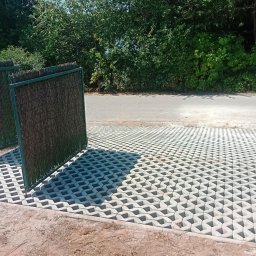 Pro-kop - Ułożona szara ażurowa kostka brukowa na podjeździe z zielonymi panelami ogrodzeniowymi. Częściowo ułożona kostka i sterta kostki brukowej w tle.