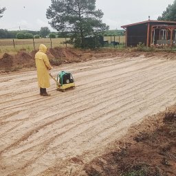 Pro-kop - Pracownik w żółtym płaszczu zagęszcza piasek zagęszczarką wibracyjną na działce budowlanej. Widoczne ślady po przejeździe maszyny i dom w tle.