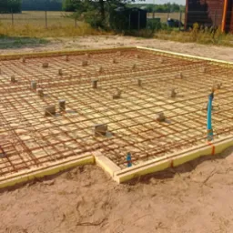 Zbrojenie fundamentu na piasku, z widocznymi podkładkami dystansowymi, szalunkiem z żółtego styropianu i wystającą rurą. W tle budynek i zieleń.