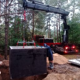 Pro-kop - Montaż betonowego szamba za pomocą dźwigu na zalesionej działce. Pracownik w ubraniu roboczym nadzoruje opuszczanie zbiornika do wykopu. Widoczny samochód ciężarowy.