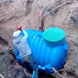 Pro-kop - Montaż niebieskiego zbiornika z zieloną pokrywą w wykopie. Pracownik w dresie i czapce zakłada pomarańczową pokrywę. Widoczne narzędzia i ziemia.