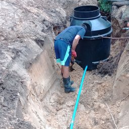 Pro-kop - Montaż szamba: mężczyzna w wykopie, w gumowych butach, instaluje zbiornik z rurą. Prace ziemne w toku, widoczne ślady kopania, naturalne oświetlenie.