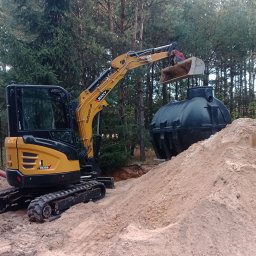 Pro-kop - Żółta mini koparka SANY obok czarnego zbiornika na tle drzew. Wykopana ziemia tworzy spory nasyp. Widoczny etap instalacji zbiornika.