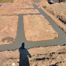 Pro-kop - Wylane fundamenty pod budowę domu, widoczne wykopane rowy zalane betonem. W tle hałda ziemi i betoniarka. Cień osoby w rogu kadru.