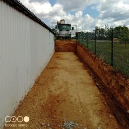 Pro-kop - Wykop fundamentu z widocznym zbrojeniem i zaparkowaną koparką SCANIA w tle, na tle błękitnego nieba z chmurami. Widok z perspektywy gruntu.