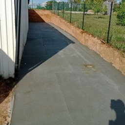 Pro-kop - Świeżo wylana, gładka betonowa droga prowadząca wzdłuż budynku z blachy trapezowej, ogrodzona siatką. Widoczny wykopany grunt po bokach.