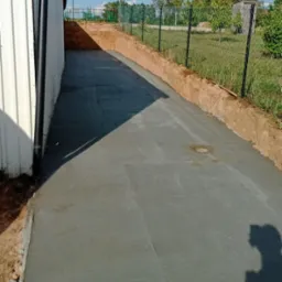 Świeżo wylana, gładka betonowa droga prowadząca wzdłuż budynku z blachy trapezowej, ogrodzona siatką. Widoczny wykopany grunt po bokach.