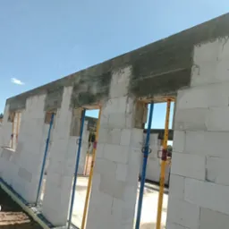 Ściana domu z betonu komórkowego w trakcie budowy, z widocznymi otworami okiennymi i betonowym wieńcem, podparta stemplami budowlanymi. Błękitne niebo w tle.