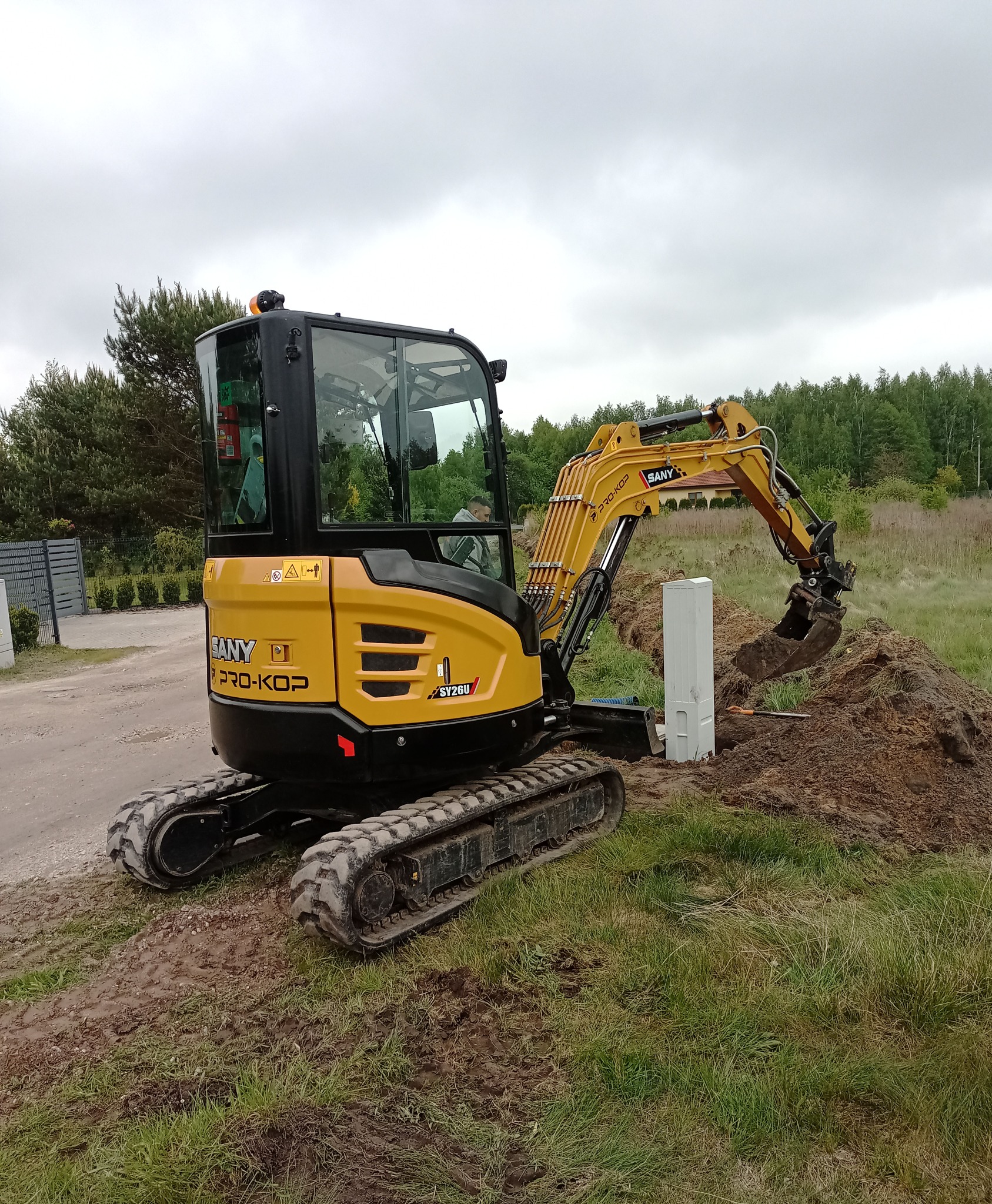 Żółta mini koparka SANY PRO-KOP SY26U wykopuje dół obok białego słupka na trawiastym terenie z drzewami w tle, przy pochmurnym niebie.