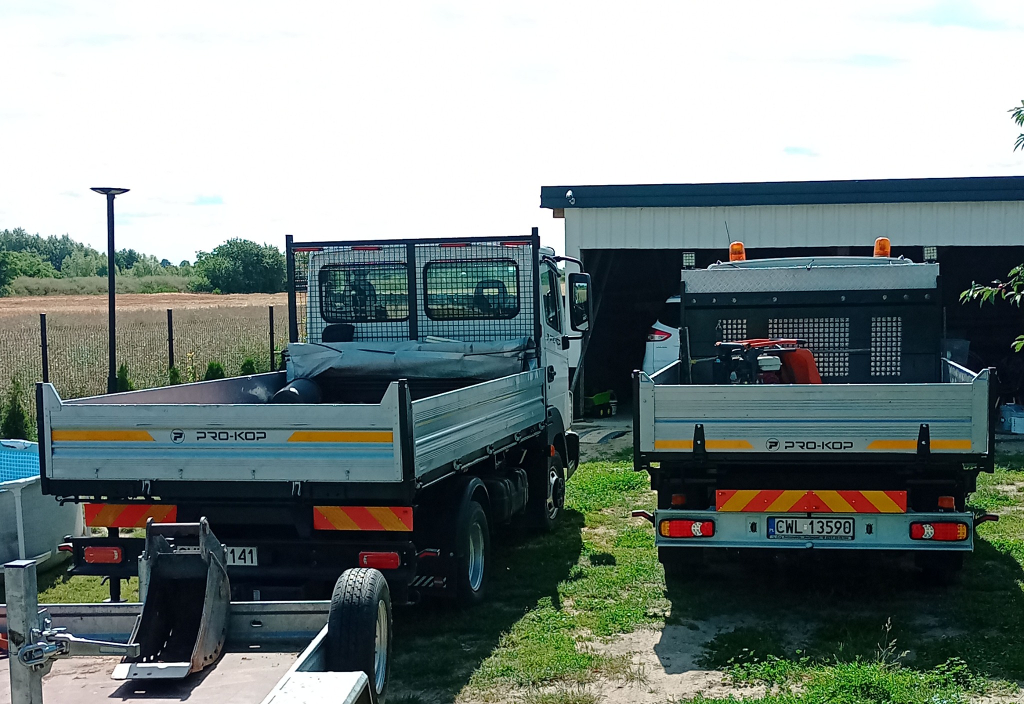 Dwa samochody ciężarowe z otwartą skrzynią ładunkową, jeden z przyczepą i łyżką koparki, na trawiastym terenie, w tle budynek gospodarczy i pola.