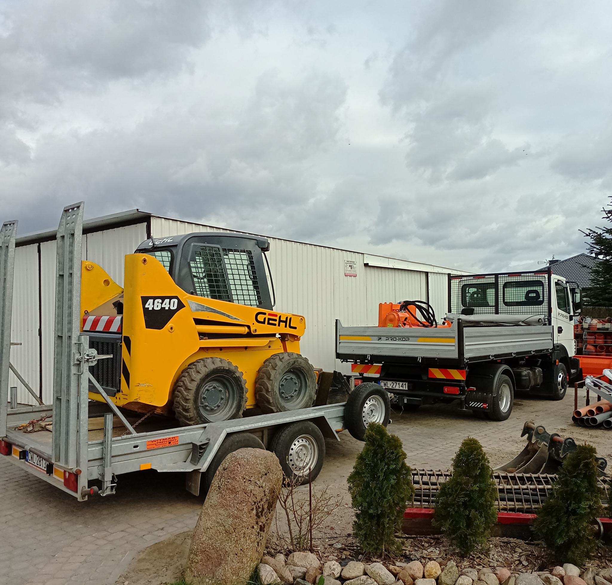 Żółta ładowarka Gehl 4640 na srebrnej przyczepie, obok biała ciężarówka Pro-Kop na tle białego budynku. Elementy krajobrazu: kamienie, tuje, pochmurne niebo.