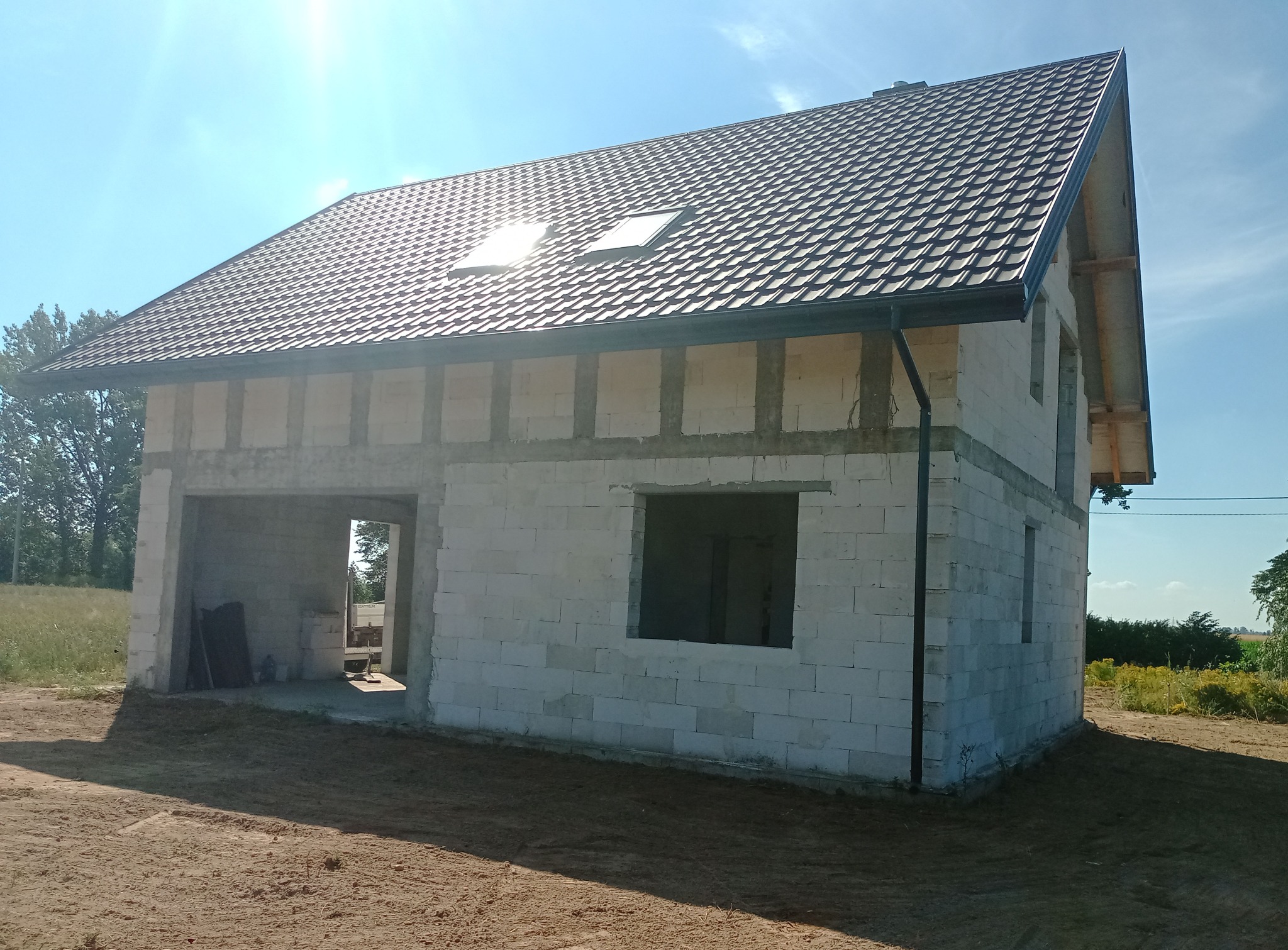 Surowy dom z białych pustaków z ciemną dachówką i dwoma oknami dachowymi, w otoczeniu zieleni i błękitnego nieba. Widoczne fundamenty i częściowo otwarte okna.