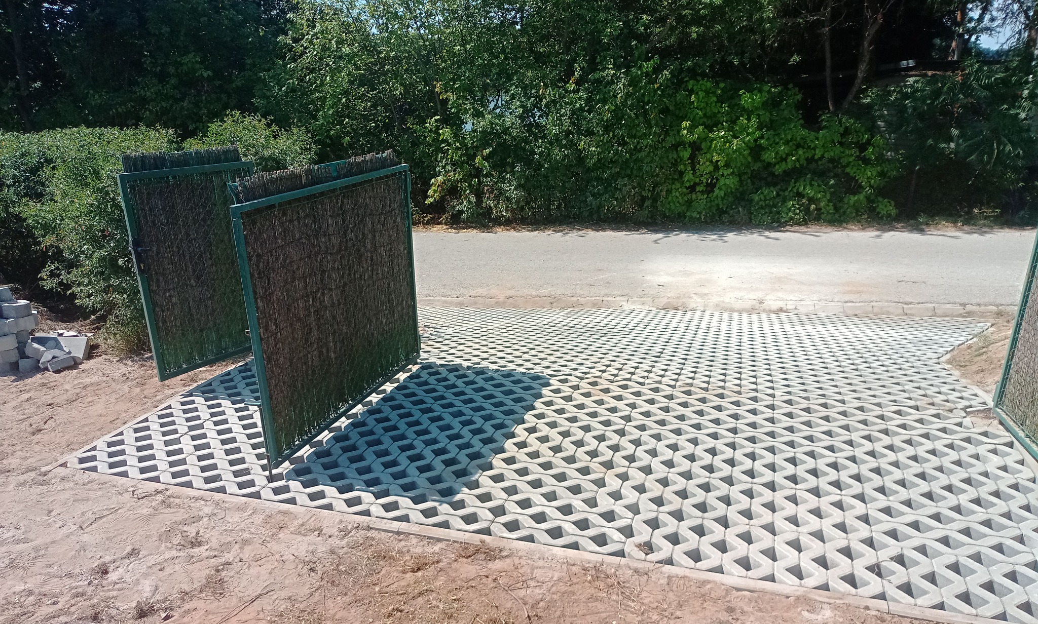 Ułożona szara ażurowa kostka brukowa na podjeździe z zielonymi panelami ogrodzeniowymi. Częściowo ułożona kostka i sterta kostki brukowej w tle.